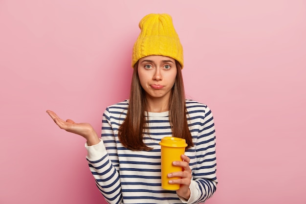 Foto di una donna europea esitante con espressione confusa, alza il palmo, tiene il caffè da asporto, indossa un cappello giallo e un maglione a righe, posa su sfondo rosa.