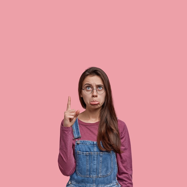 Foto di una donna scontenta con i capelli lunghi e scuri, si sente stressata dalle cattive notizie, indossa occhiali da vista e tute di jeans