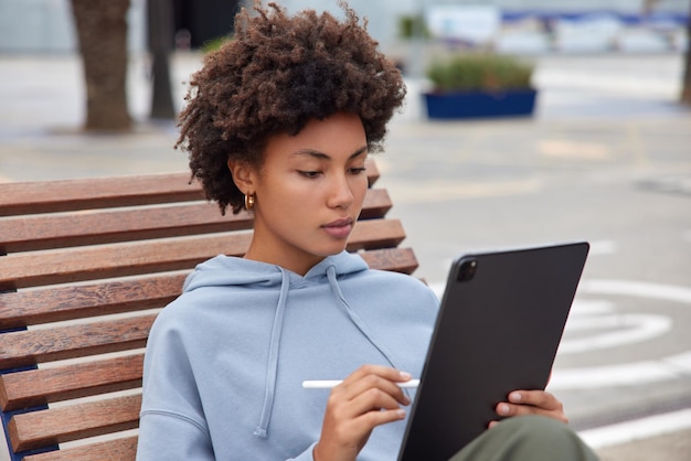 Foto di una donna seria concentrata sullo schermo del tablet disegna immagini con lo stilo in posa su una panca di legno vestita con una felpa con cappuccio che trascorre il tempo libero all'aperto su sfondo sfocato lavora come freelance