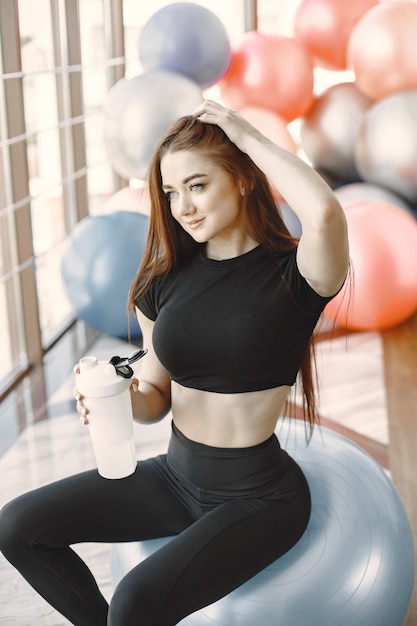Foto di una donna sportiva sorridente in posa con una bottiglia d'acqua. Indossa abiti sportivi scuri. Seduto su una palla in forma.
