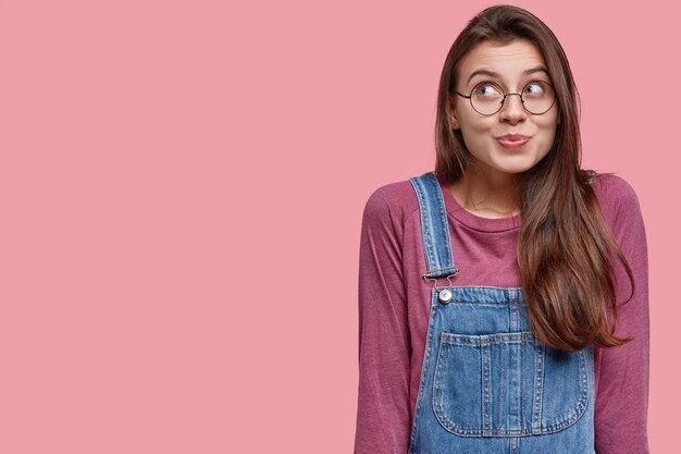 Foto di una giovane donna sognante con i capelli lunghi, indossa occhiali rotondi, guarda felicemente in alto, ha occhiali rotondi, tuta in denim