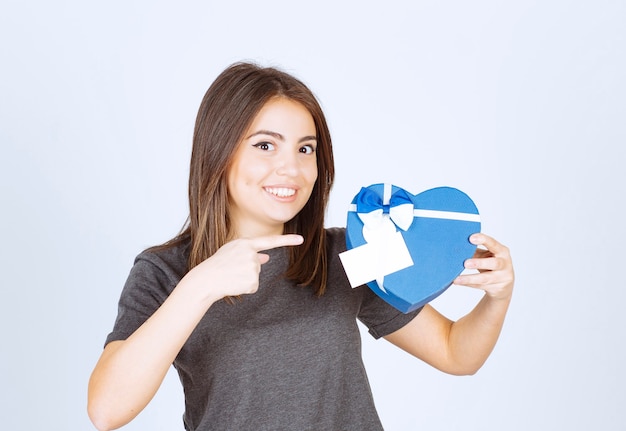 Foto di una giovane donna sorridente che indica una confezione regalo a forma di cuore.