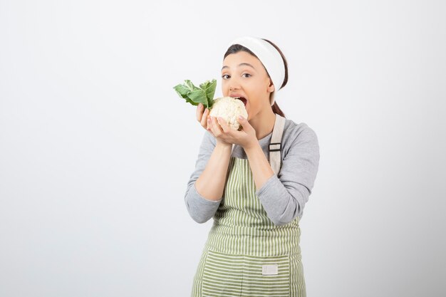 Foto di una giovane modella simpatica in grembiule che mangia un cavolfiore