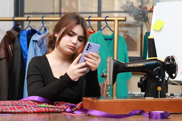 Foto di una giovane sarta seduta alla scrivania e guardando il suo telefono