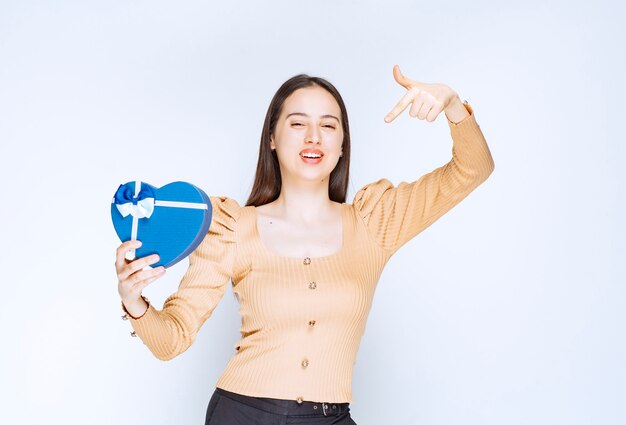 Foto di una modella di giovane donna che indica una scatola regalo a forma di cuore contro il muro bianco.