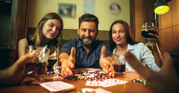 Foto di vista laterale di amici maschi e femmine seduti al tavolo di legno. Uomini e donne che giocano a carte. Mani con il primo piano dell'alcool. Poker, intrattenimento serale e concetto di eccitazione