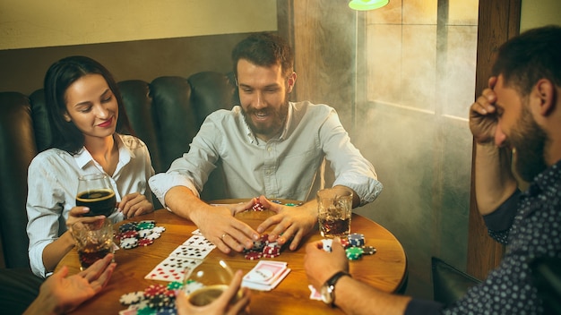 Foto di vista laterale di amici maschi e femmine seduti al tavolo di legno. Uomini e donne che giocano a carte. Mani con il primo piano dell'alcool.
