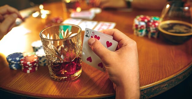 Foto di vista laterale di amici maschi e femmine seduti al tavolo di legno. Uomini e donne che giocano a carte. Mani con il primo piano dell'alcool.