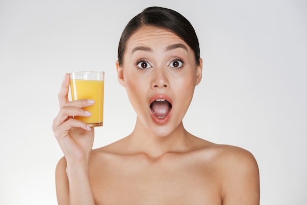 Foto divertente della donna divertente con capelli scuri in panino che tiene vetro trasparente di succo d'arancia appena spremuto, isolato sopra la parete bianca