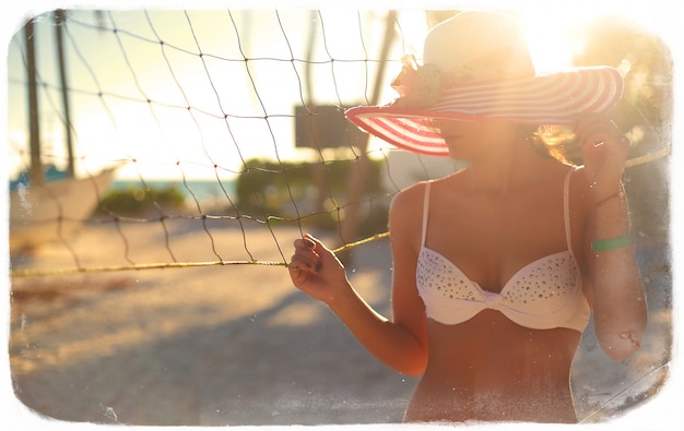 Foto in stile retrò di ragazza modello sexy in bikini bianco con rete da pallavolo sulla spiaggia e palme dietro il cielo estivo blu