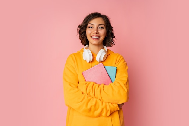 Foto in studio di studente carino felice con quaderni e auricolari in piedi sul rosa.