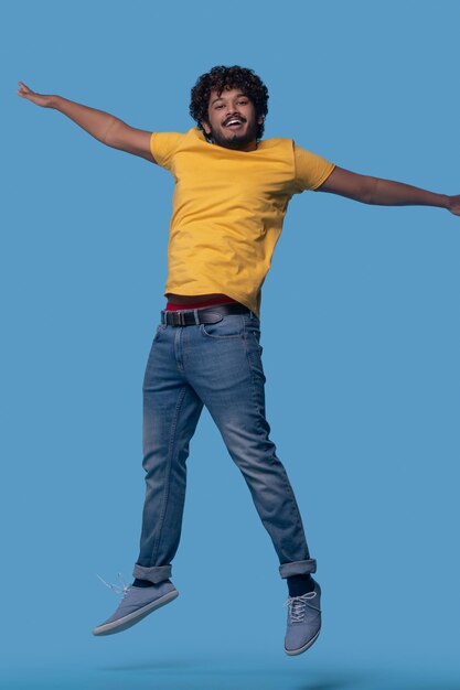 Foto in studio di un maschio indiano dai capelli ricci sorridente e felice che salta sullo sfondo blu