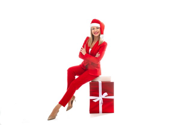 Foto in studio di una donna d'affari elegante e attraente in abito rosso brillante, tacchi e cappello da Babbo Natale in posa su un regalo di Natale avvolto in carta rossa e legato con fiocco. Isolare su bianco.