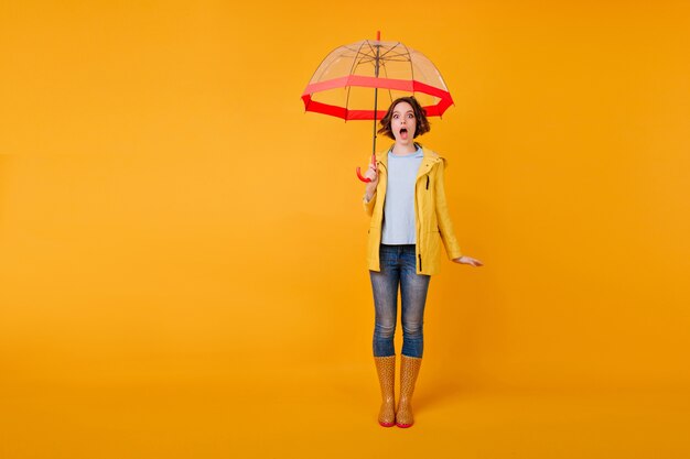 Foto integrale della ragazza scioccata con la bocca aperta in piedi con l'ombrello. Giovane signora alla moda in blue jeans in posa con l'espressione del viso stupito in studio.