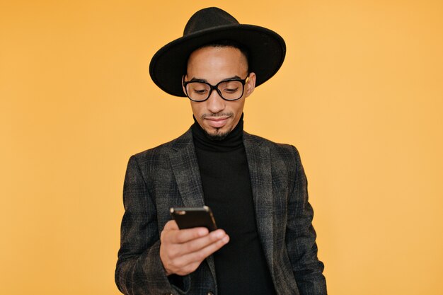 Foto interna del giovane nero in abbigliamento splendido che tiene smartphone. Ragazzo africano emotivo in attesa di chiamata con un sorriso.