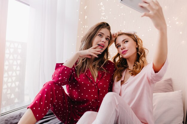 Foto interna dell'adorabile ragazza bionda che utilizza il telefono per selfie con un'amica. Entusiasta signora bruna in abito da notte di cotone rosso in posa con la sorella.