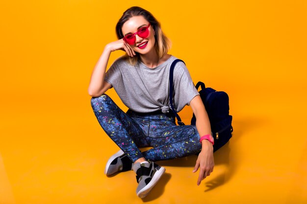 Foto interna della ragazza in leggings colorati e camicia alla moda seduta sul pavimento. Ritratto dello studio di giovane donna alla moda in scarpe sportive in posa su sfondo giallo e tenendo lo zaino.