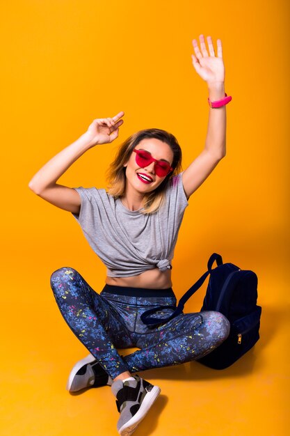 Foto interna della ragazza in leggings colorati e camicia alla moda seduta sul pavimento. Ritratto dello studio di giovane donna alla moda in scarpe sportive in posa su sfondo giallo e tenendo lo zaino.