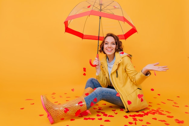 Foto interna di una bella ragazza in abbigliamento autunnale che ride mentre posa sul pavimento con l'ombrellone. Carina donna riccia in jeans godendo il servizio fotografico nel giorno di San Valentino.