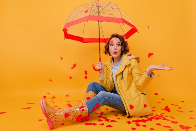 Foto interna di una ragazza spettacolare che indossa scarpe di gomma e pantaloni di jeans blu in posa con l'ombrello. Ritratto di gioiosa signora seduta sul pavimento con cuori di carta rossa.