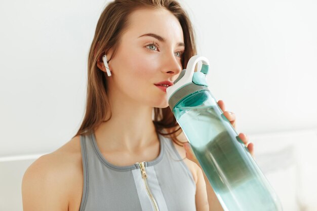 Foto ravvicinata della giovane donna in top sportivo che guarda sognante da parte con una bottiglia d'acqua e auricolari mentre si è in piedi in cucina