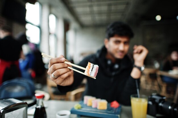 Foto ravvicinata di bastoncini che tengono un giovane asiatico casual ed elegante con gli auricolari al bar che mangia sushi