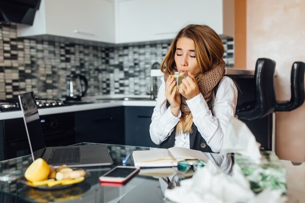 Foto ravvicinata, giovane donna malata con una sciarpa calda seduta sul tavolo in cucina, tiene una tazza con il tè