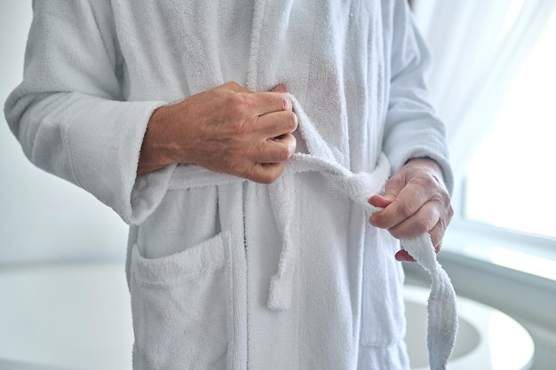 Foto ritagliata di un maschio in piedi vicino alla vasca da bagno mentre si lega la cintura del suo accappatoio in spugna