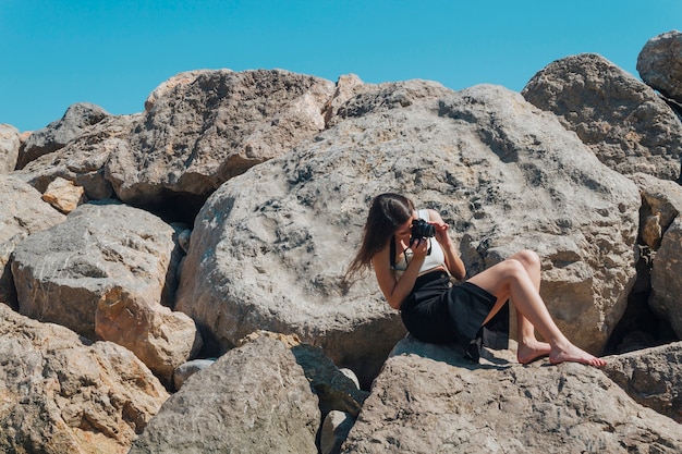 Fotografo femminile che si siede sulla roccia che prende foto con la macchina fotografica vicino al mare