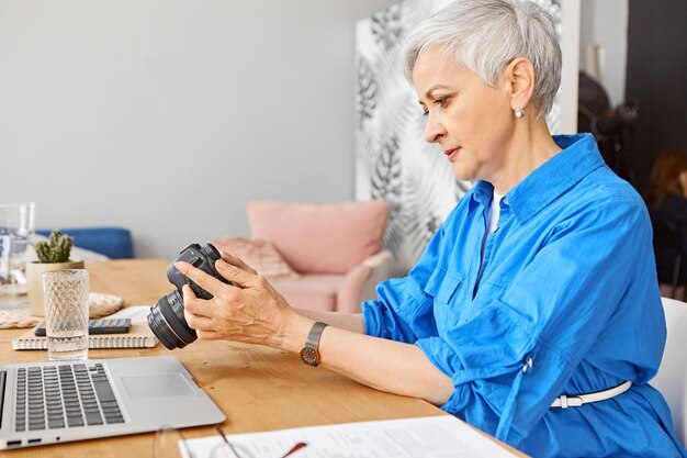 Fotografo professionista concentrato della donna matura che controlla le anteprime sulla macchina fotografica. Grave donna in pensione guardando tutorial sulla fotografia online utilizzando laptop. Hobby, lavoro a distanza e concetto di età