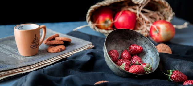 Fragole dentro il canestro della ciotola, dei biscotti, della tazza e della mela su una stuoia nera.