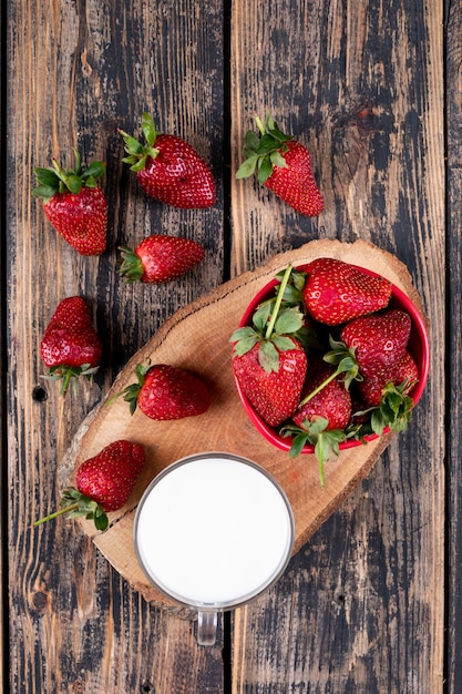 fragole in ciotola e intorno con una tazza di latte sul troncone e tavolo in legno.