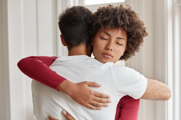 Fratello e sorella amichevoli di razza mista si abbracciano, si sostengono in situazioni di vita difficili, posano al chiuso, esprimono buone relazioni, esprimono amore. Coppia romantica interrazziale si incontra a casa