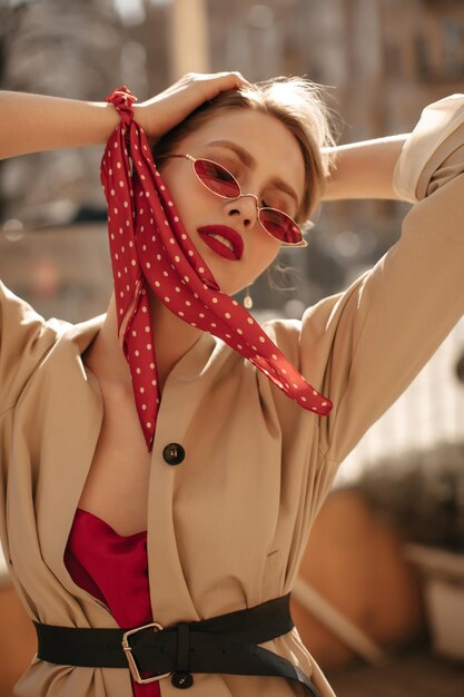 Fresca ed elegante signora bionda con labbra rosse in occhiali da sole luminosi camicetta di seta e trench beige guarda in basso e posa fuori