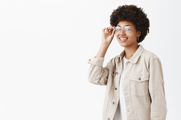 fresca, sicura ed elegante donna da ragazzo con acconciatura afro che tocca il bordo degli occhiali sugli occhi e sorride ampiamente essendo sicura di sé e pronta a sfidare la nuova giornata lavorativa