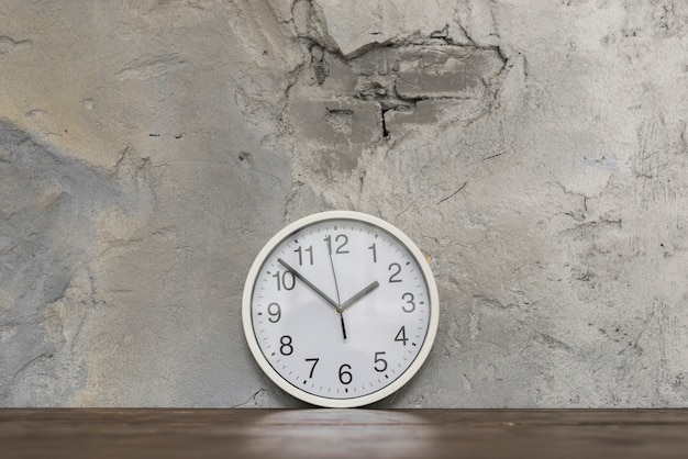 Fronte di orologio rotondo che si appoggia contro il muro di cemento nocivo sullo scrittorio di legno