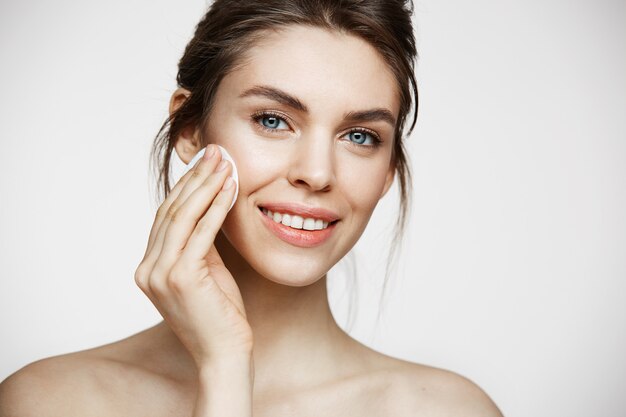 Fronte di pulizia della bella ragazza castana naturale sveglia con la spugna di cotone che sorride esaminando macchina fotografica sopra fondo bianco. Cosmetologia e spa.