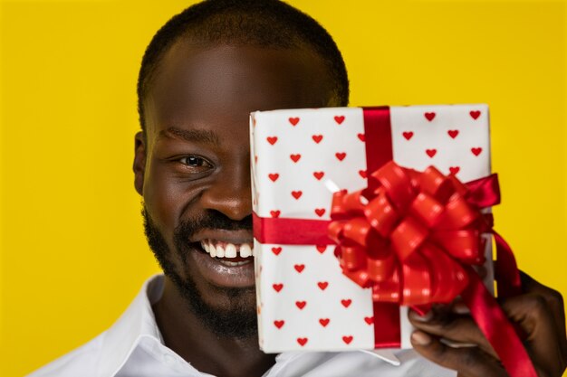Frontview di ridere giovane ragazzo afroamericano barbuto con un regalo in mano che ha chiuso metà del viso in camicia bianca