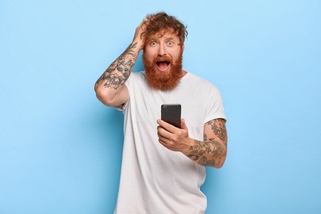 Frustrato e stressante hipster allo zenzero tiene la mano sulla testa, guarda con espressione facciale preoccupata, apre la bocca, tiene il cellulare moderno, prova paura per aver commesso un errore, ha fatto qualcosa di sbagliato con l'app