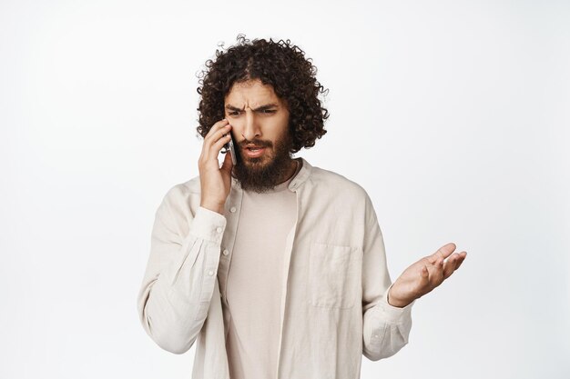 Frustrato giovane uomo mediorientale parlando al telefono cellulare con faccia confusa scrollando le spalle sconvolto in piedi su sfondo bianco