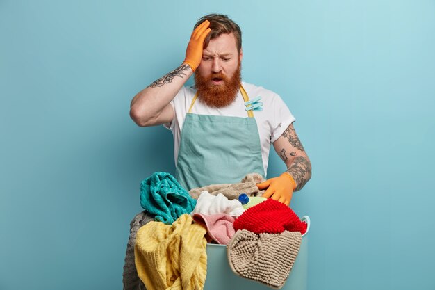 Frustrato, sopraffatto, perplesso, l'uomo volubile ha molto lavoro per la casa, tiene la mano sulla testa e fissa il cesto pieno di biancheria, ha tempo di lavarsi a casa, non sa come iniziare, vestito con il grembiule