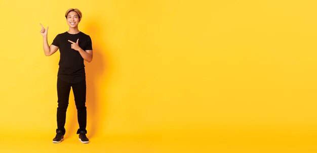 Fulllength di sorridente attraente ragazzo coreano che punta le dita a sinistra mostrando logo sfondo giallo