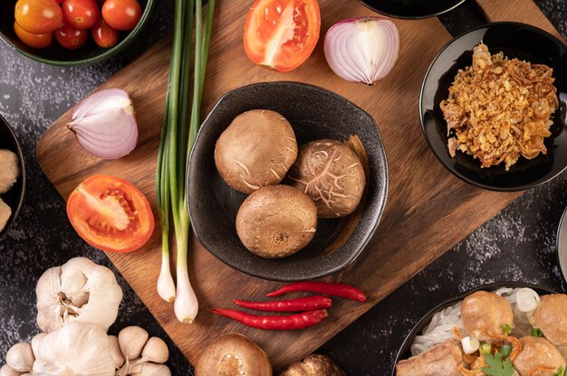 Funghi shiitake con aglio, pomodoro, peperone, cipollotto e cipolle rosse su un pavimento di cemento nero.