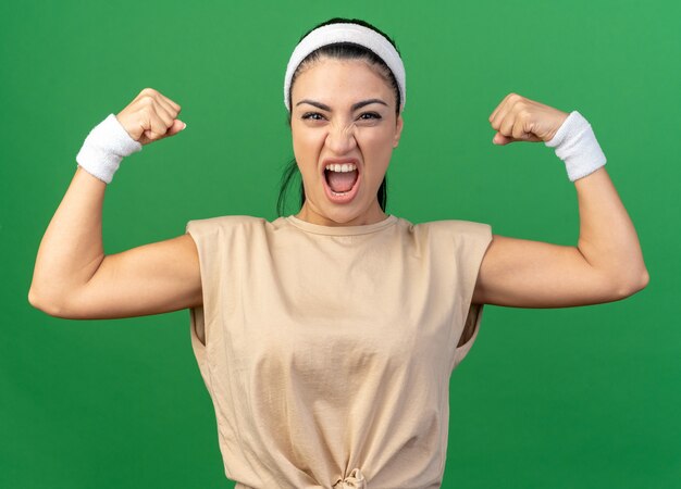 Furiosa giovane ragazza sportiva caucasica che indossa fascia e braccialetti facendo un gesto forte urlando isolata sul muro verde