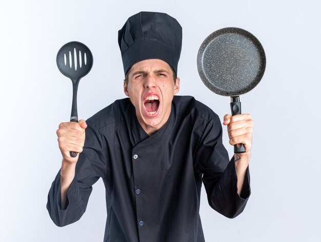 Furioso giovane cuoco maschio biondo in uniforme da chef e berretto che guarda l'obbiettivo che mostra spatola e padella urlando isolato sul muro bianco