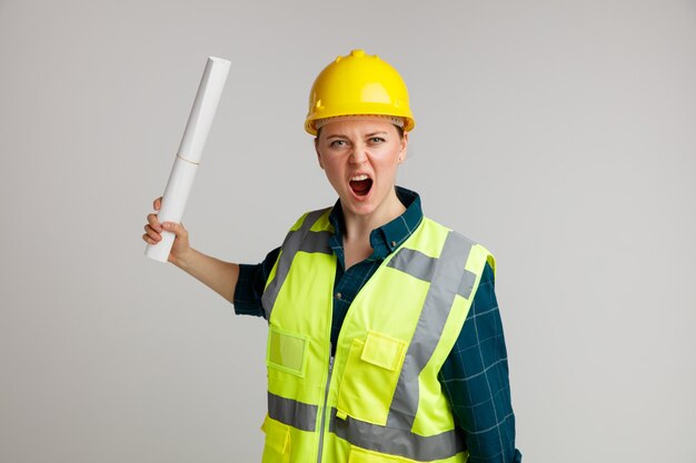 Furioso giovane operaio edile femminile che indossa il casco di sicurezza e giubbotto di sicurezza che tiene carta gridando ad alta voce