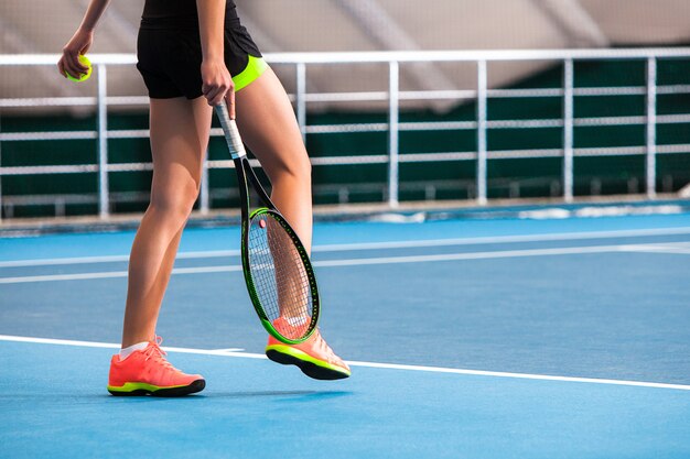 Gambe della ragazza in un campo da tennis chiuso con palla e racchetta