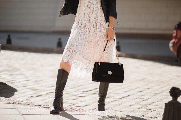 Gambe di giovane donna graziosa in stivali che camminano in strada in abito alla moda, tenendo la borsa, indossando giacca di pelle nera e abito di pizzo bianco, stile primavera autunno