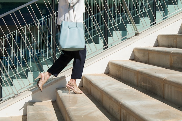 Gambe femminili in pantaloni neri con borsa blu in piedi alle scale