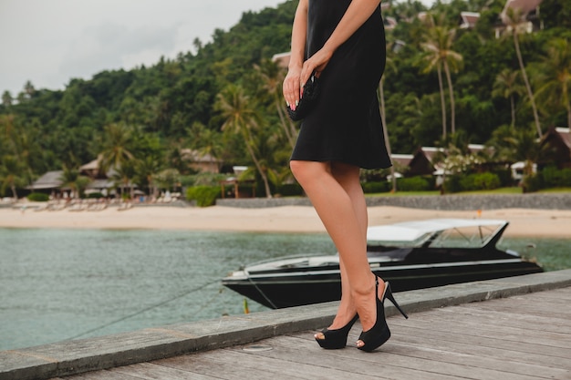 Gambe in scarpe nere col tacco alto della donna attraente sexy di lusso vestita in abito nero in posa sul molo in hotel resort di lusso, vacanze estive, spiaggia tropicale